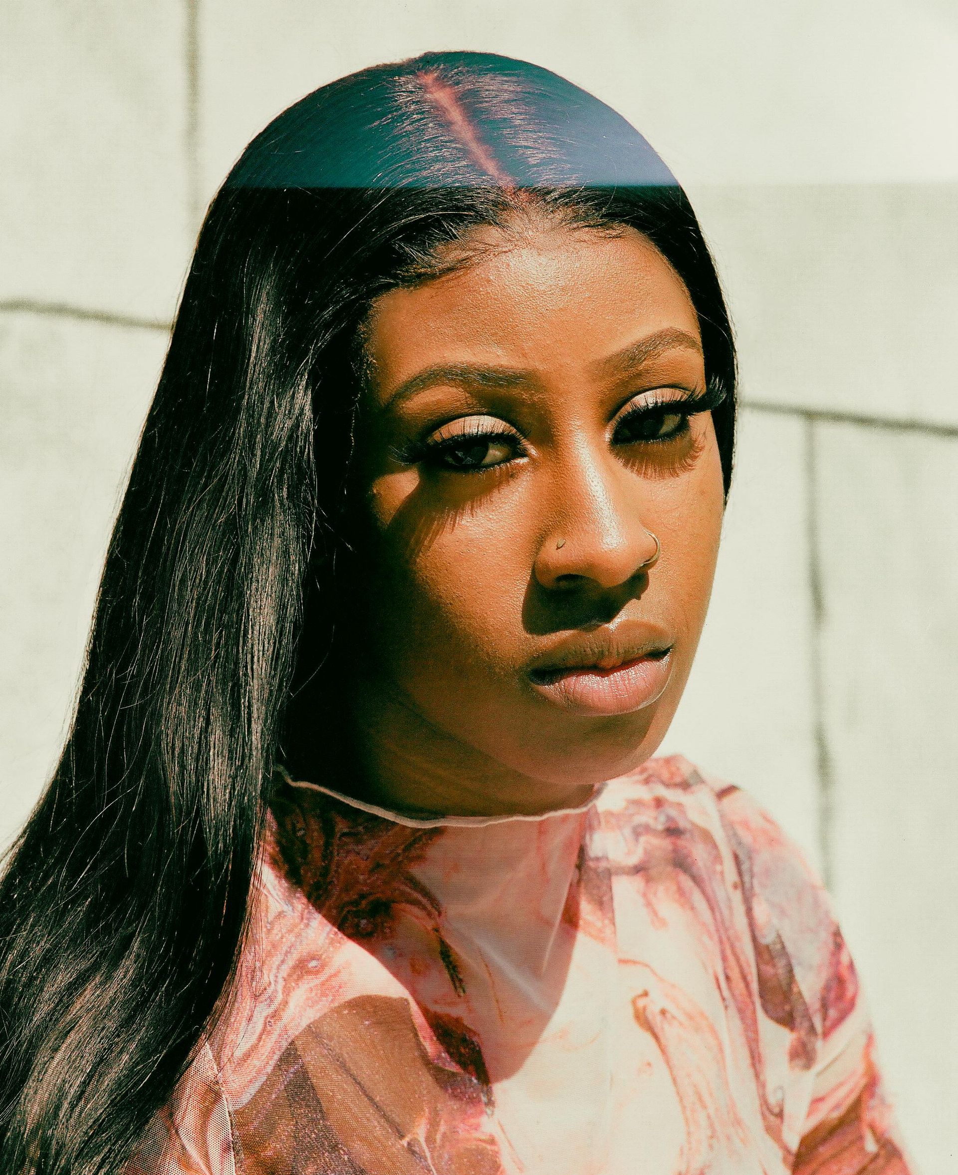 Woman with long black hair, wearing a pink patterned shirt, gazes slightly downward.