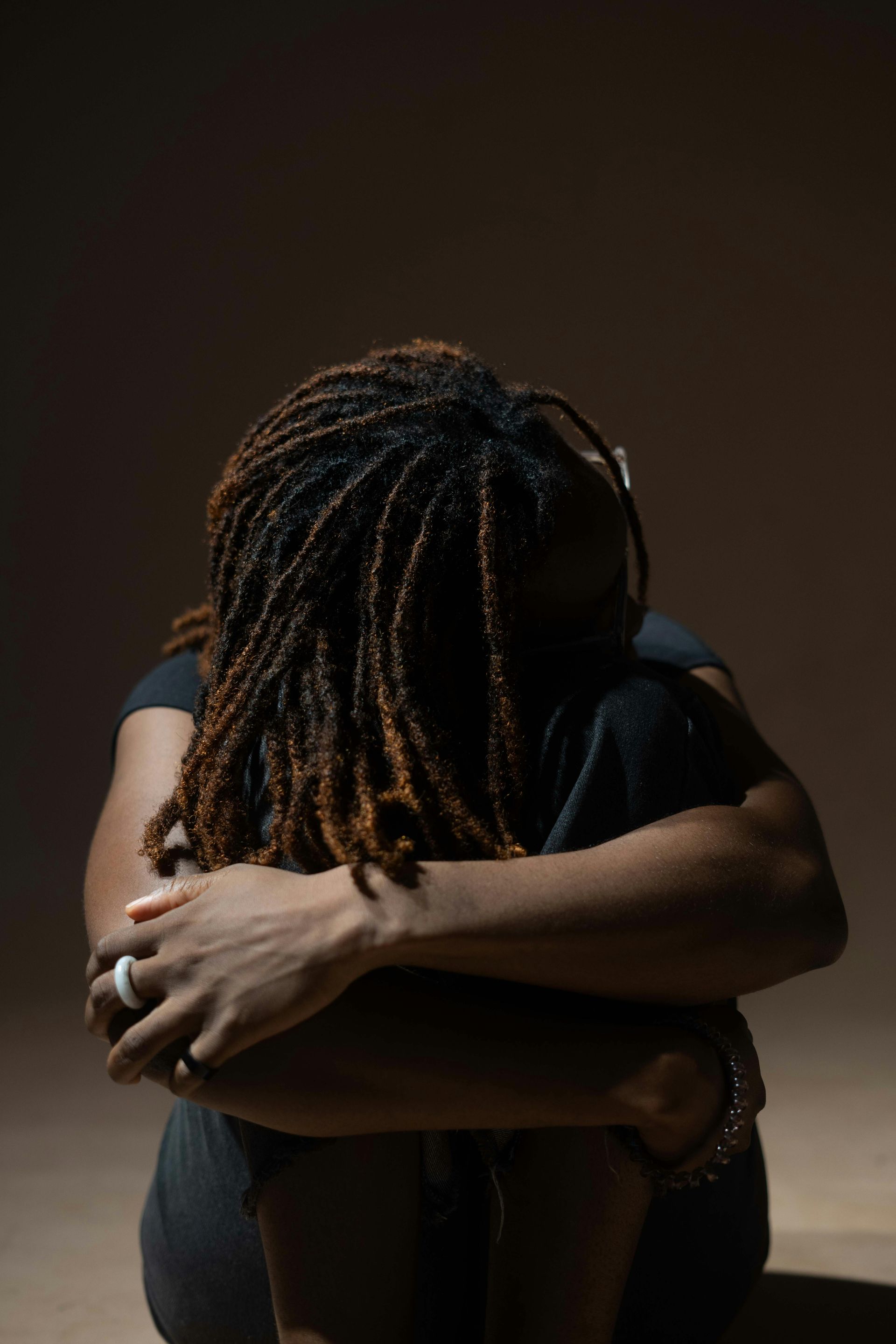 Person with dreadlocks, huddled with arms wrapped around legs. Dark setting, appearing distressed.