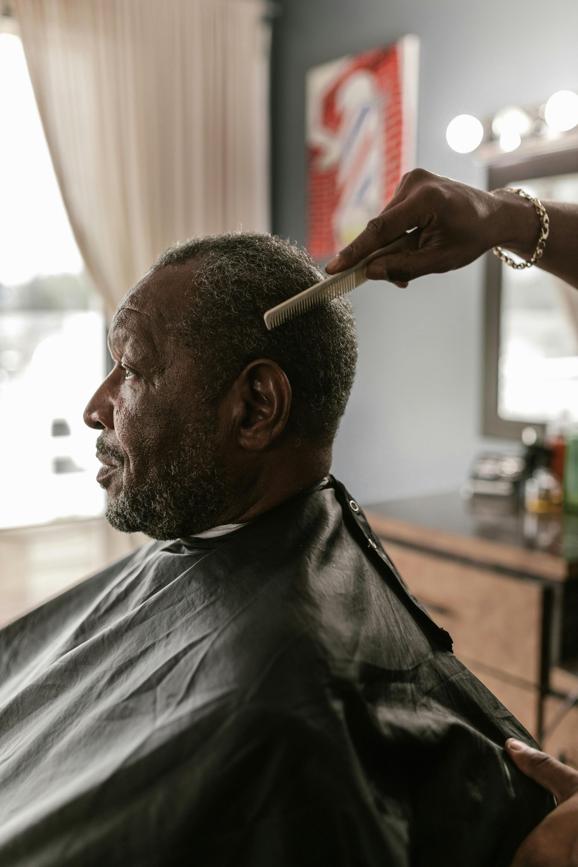 Barber combing an older Black man's hair in a barbershop.