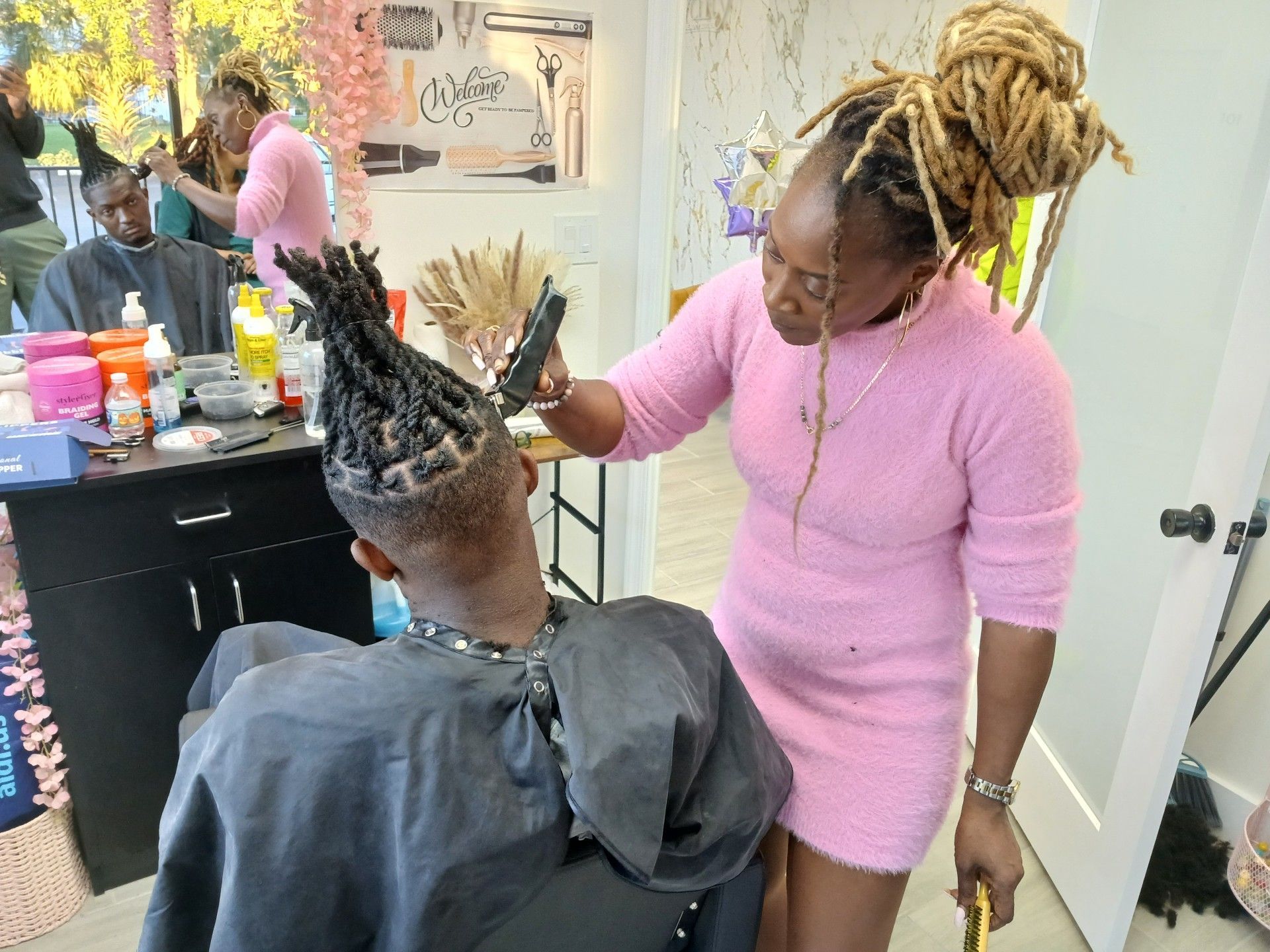 A woman in pink cuts a man's dreadlocks in a salon.