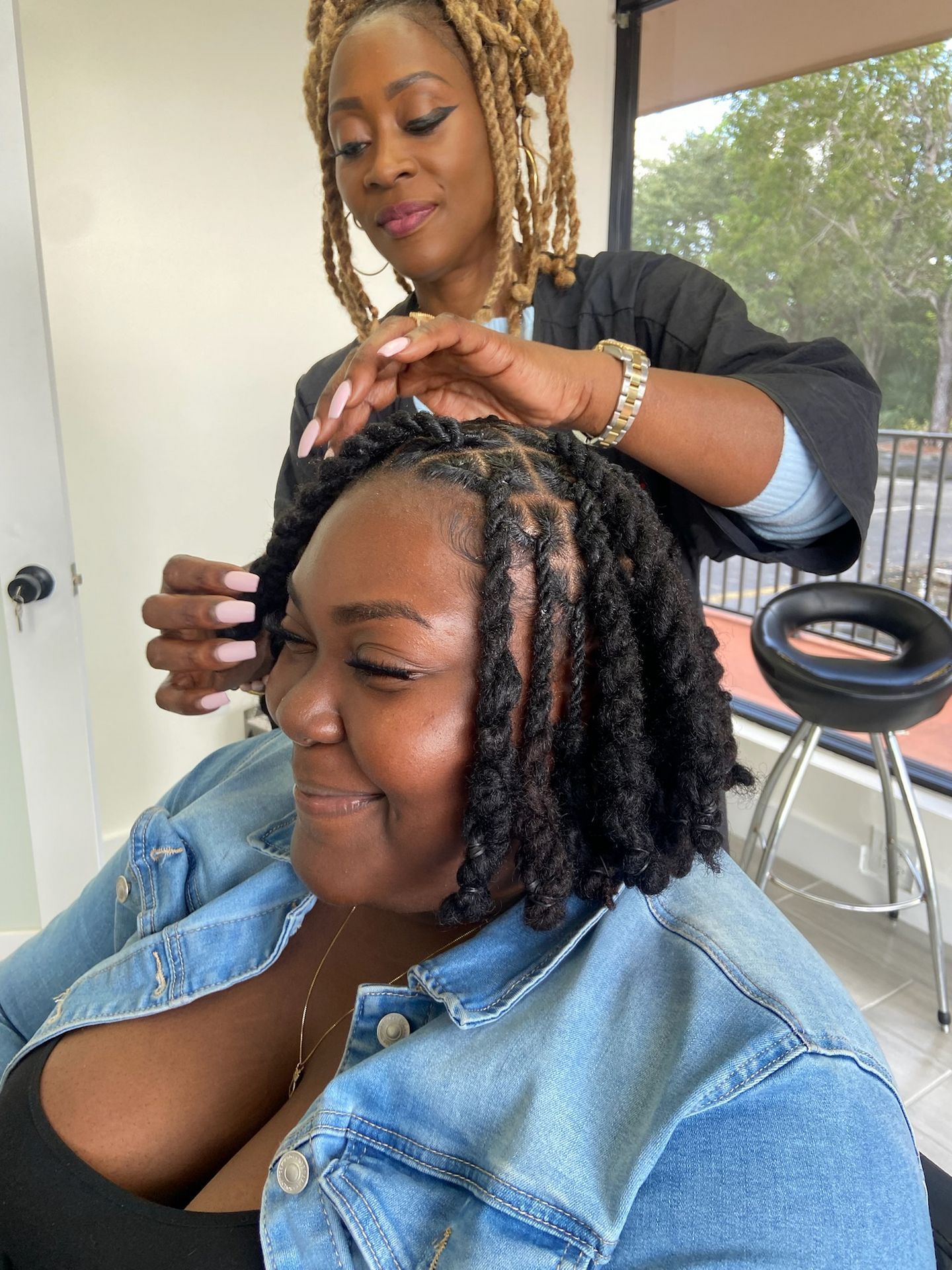 Woman getting her hair styled at a salon. Stylist with blonde locs touches her hair. Both smiling.