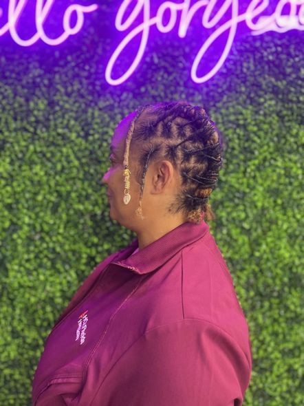 Woman with braided hair, wearing a maroon jacket, standing in front of a green wall with a neon sign.