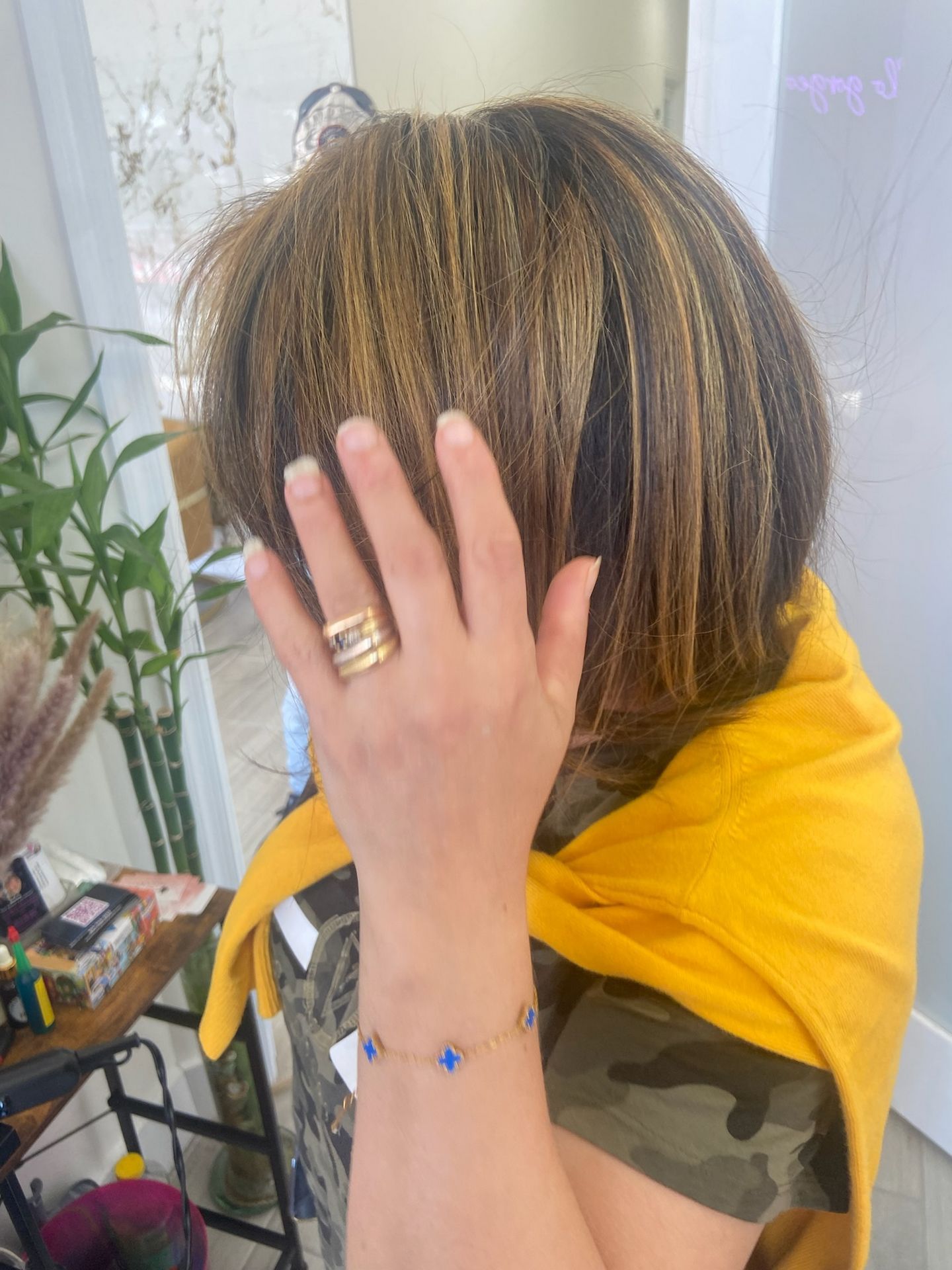 Woman with layered brown and gold hair covers face with hand, wearing gold rings and blue gemstone bracelet. Yellow sweater.