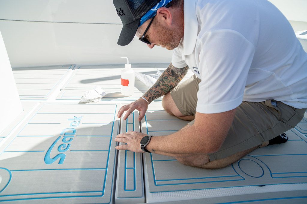 Man installing boat decking, kneeling, wearing sunglasses and a hat. Grey and blue colors.
