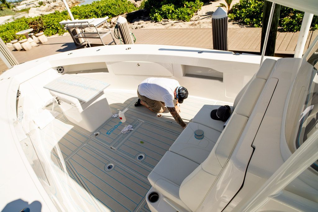 Man kneels on boat deck, cleaning. White boat with grey deck, sunny outdoors.