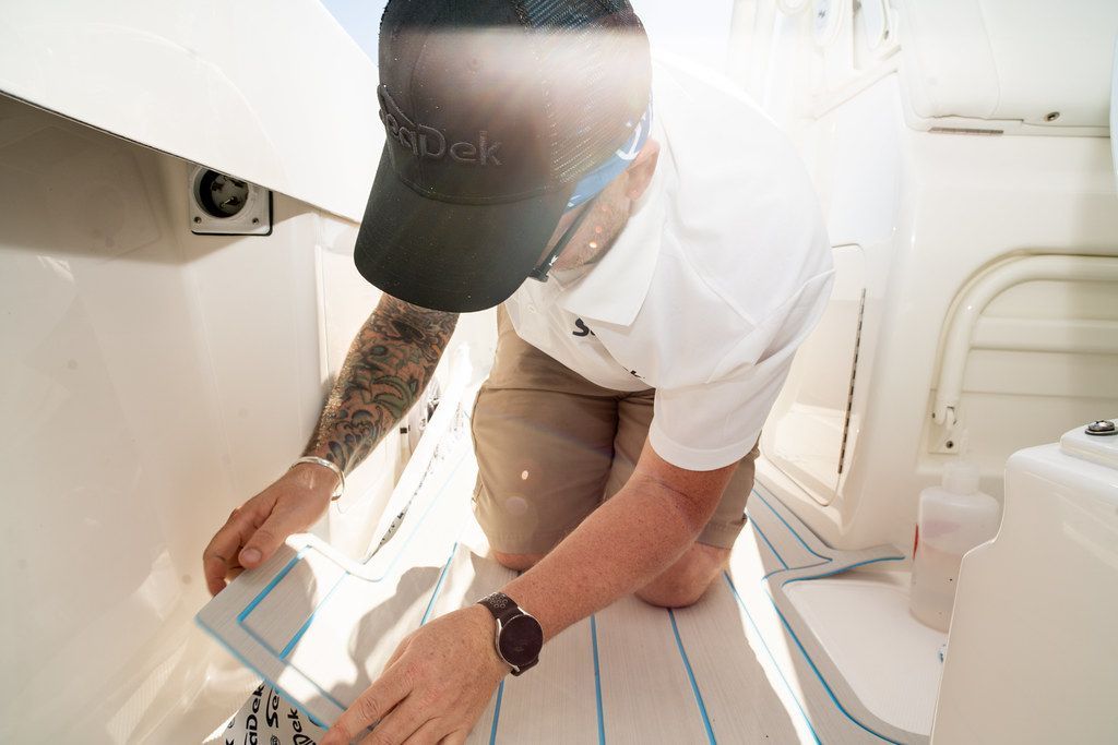 Person kneeling on a boat, installing blue-striped flooring, wearing a hat and watch.