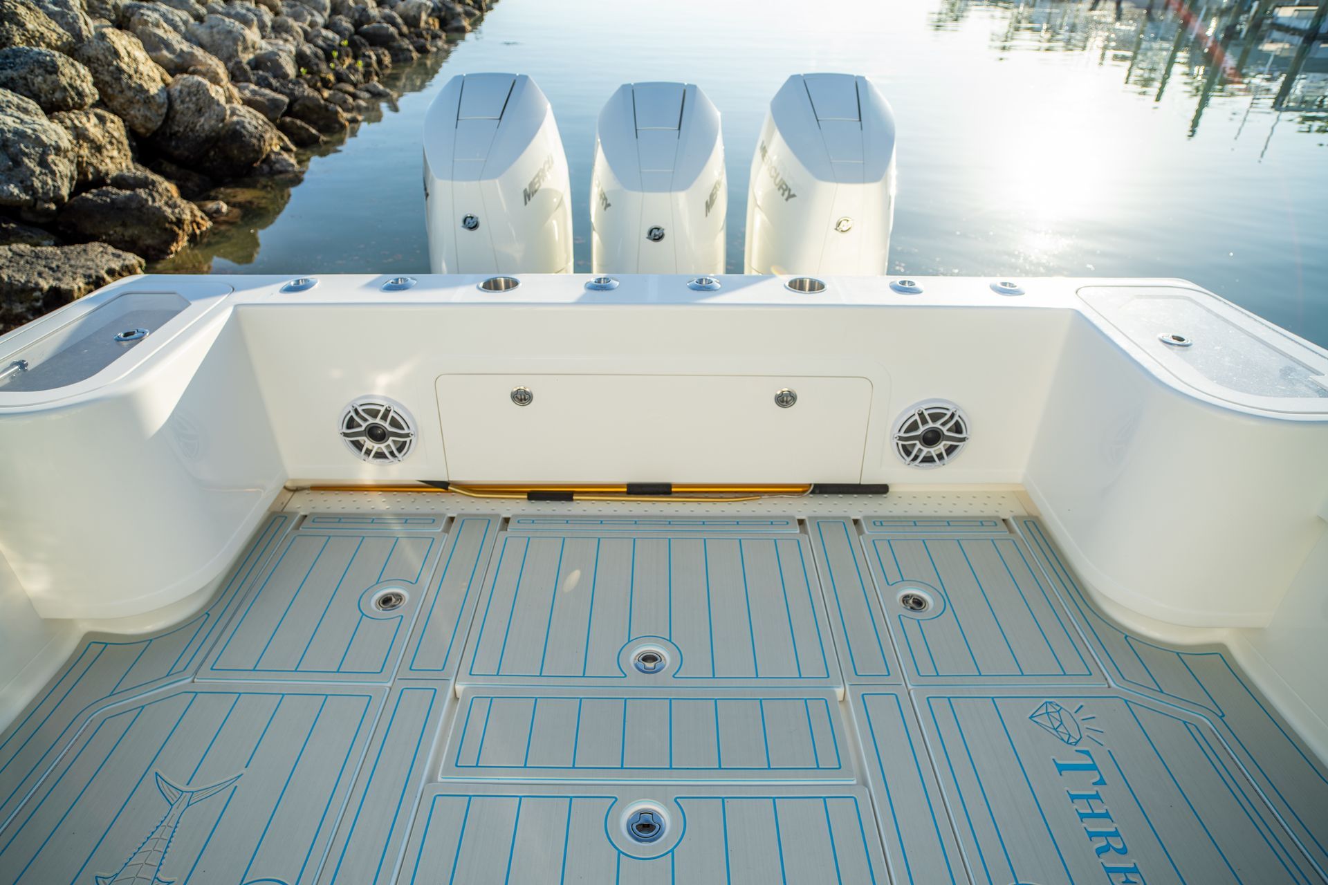 A white boat's stern with three outboard motors, speakers, and patterned flooring; setting is a sunny harbor.