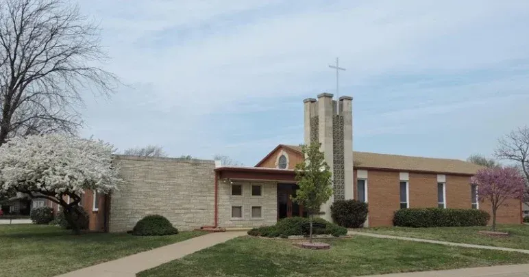 Church building with a cross on the steeple, brick and stone exterior, trees, and cloudy sky.