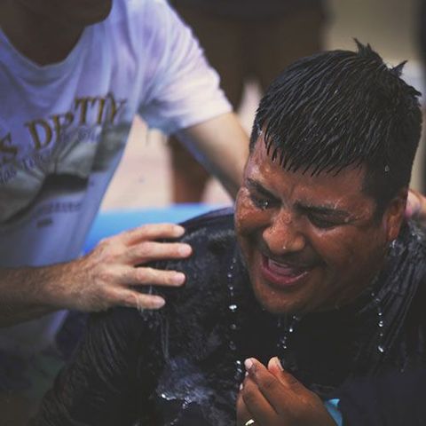 Man being baptized in a pool of water, smiling, with water droplets on his face.