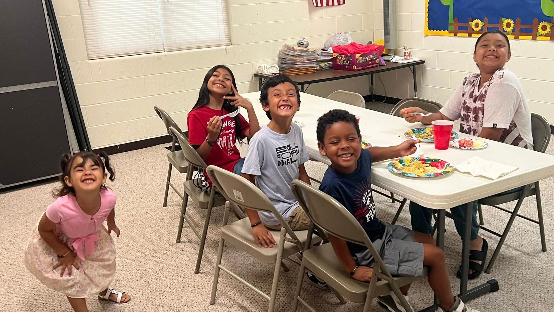 Children in a circle raising hands in a room with blue carpet and shelves.