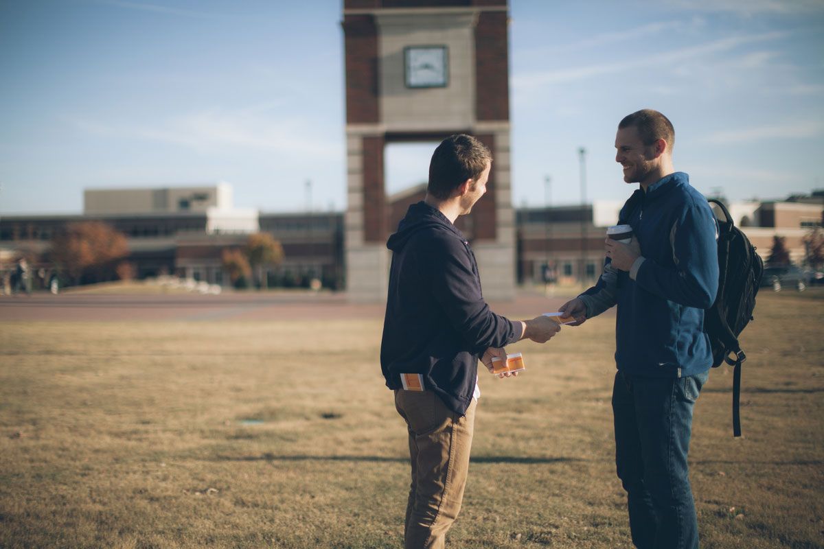 Two people shaking hands outdoors near a clock tower. Sunny day, open field, one holds a coffee cup.