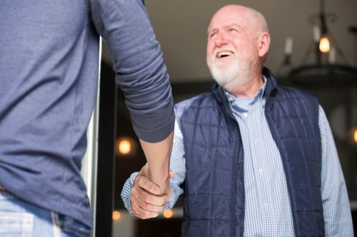 Man shaking hands with another person, both smiling; indoors, lit background.