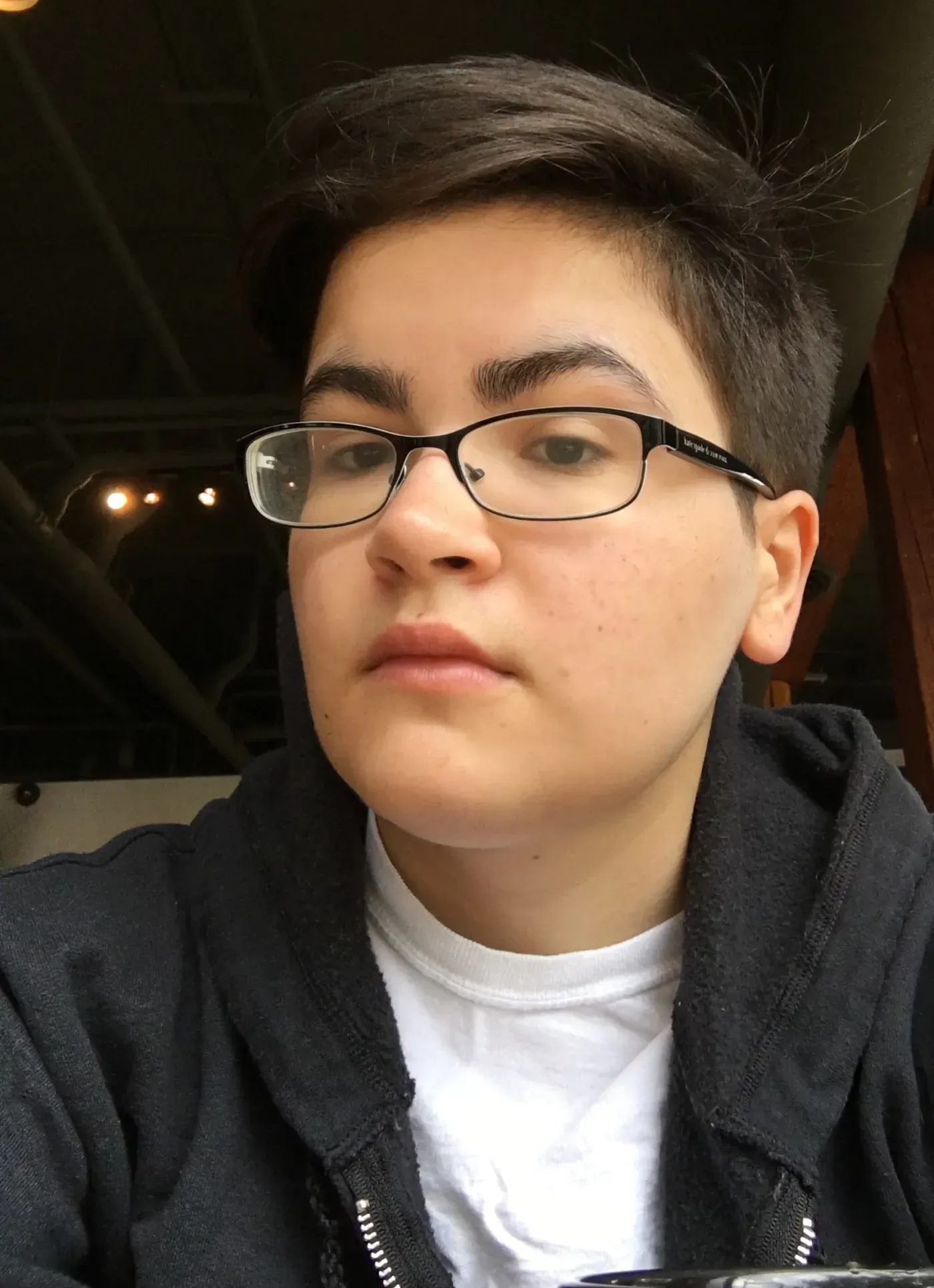 A young man wearing glasses and a black jacket is taking a selfie.