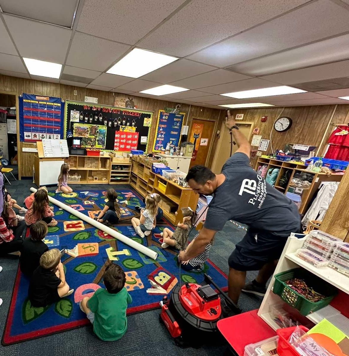 A group of children are sitting on the floor in a classroom while a man vacuums the floor.