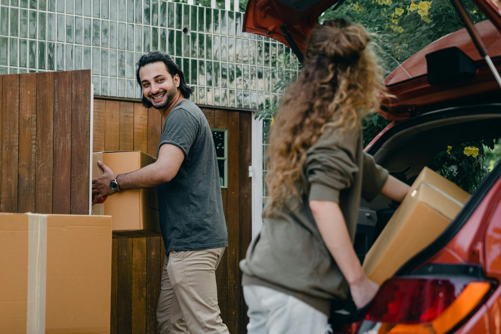 Couple moving boxes, loading them into a red car. Man smiles, holding box; woman in car's trunk. Outdoor setting.