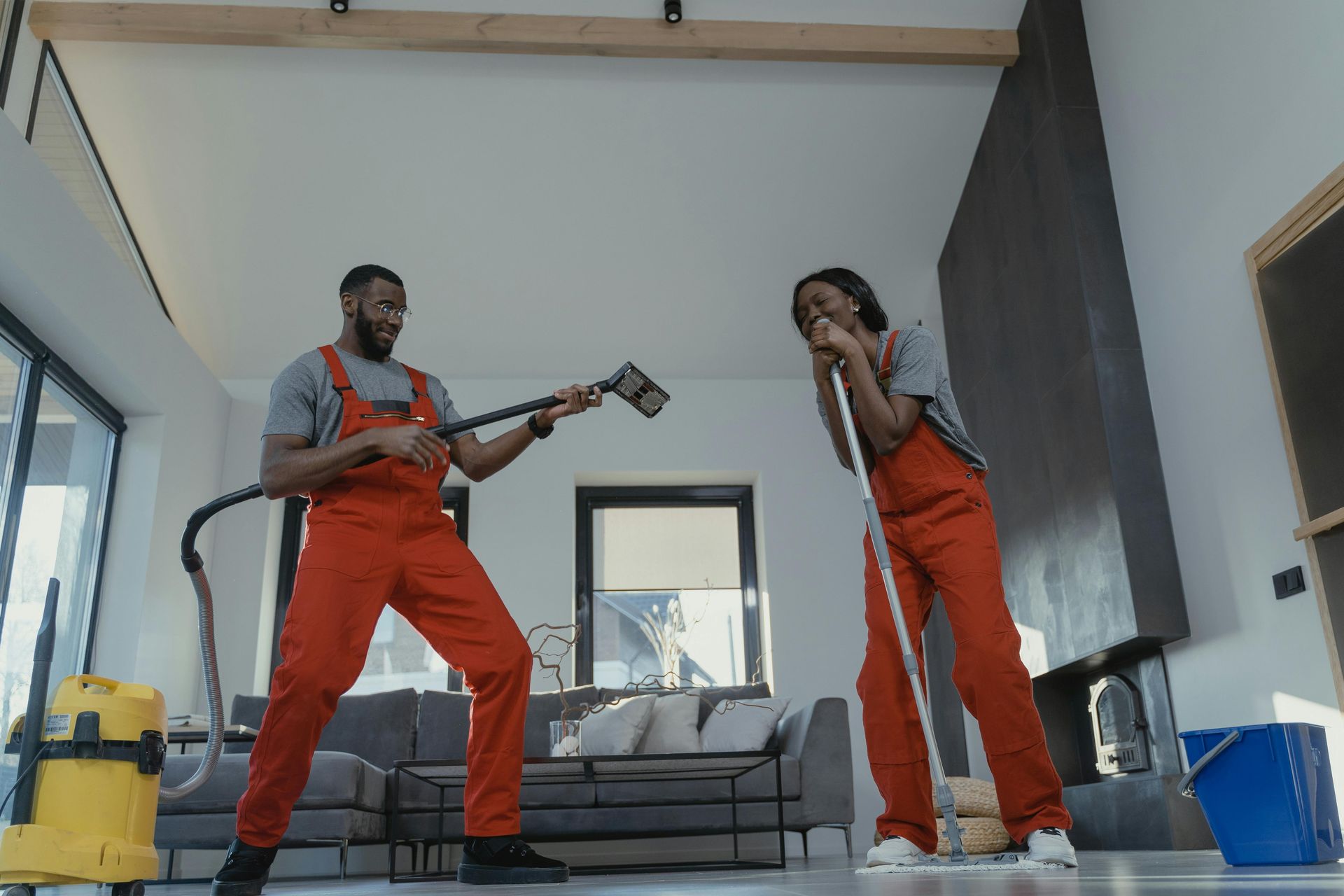 Two people in orange jumpsuits dancing with cleaning tools in a living room.
