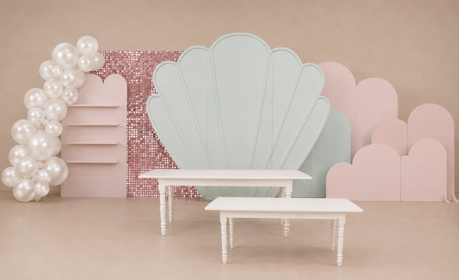 Painel de fundo para festa em tons pastel com decoração de conchas, guirlanda de pérolas e pequenas mesas brancas.