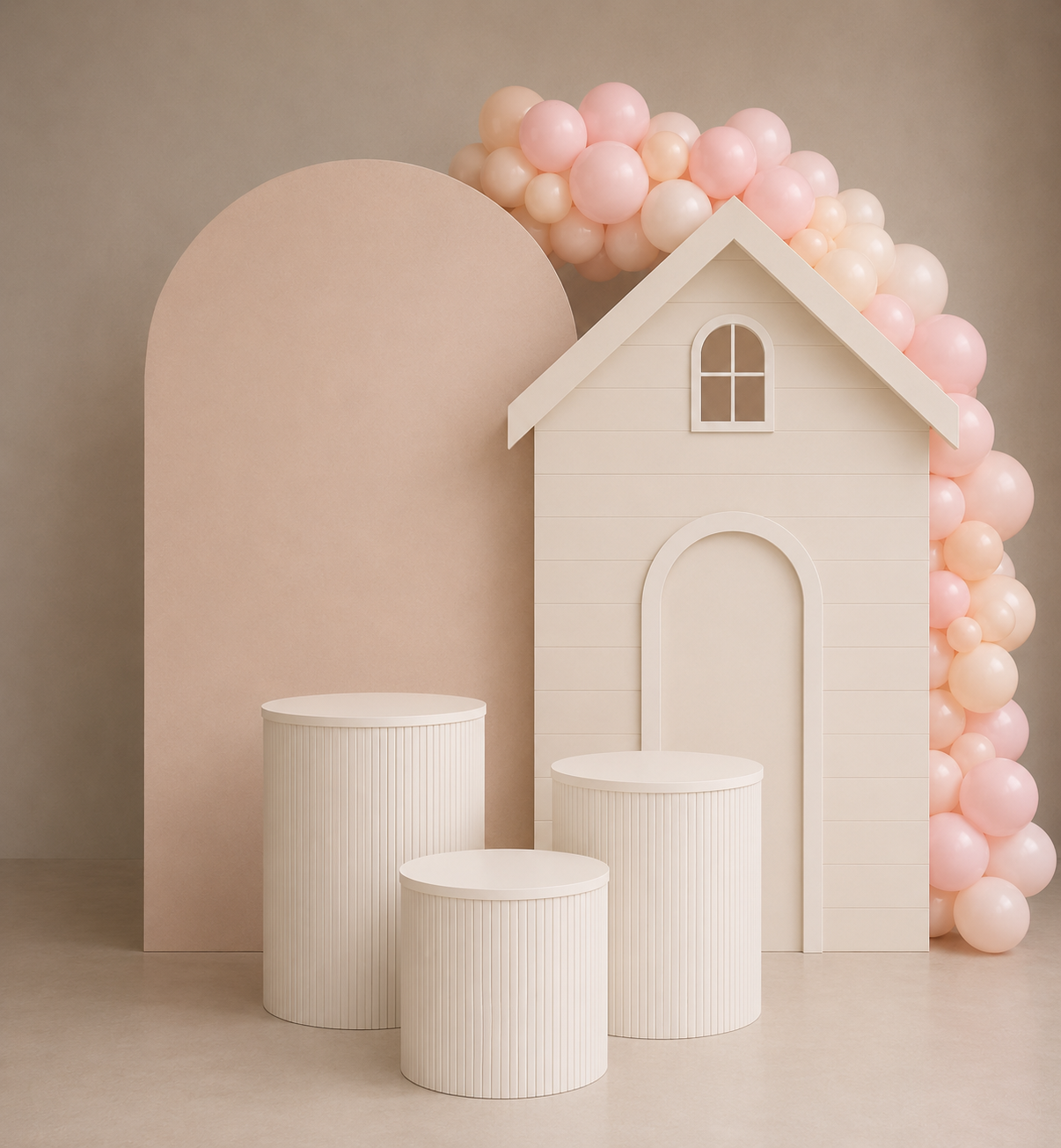 Casa cenográfica em tons pastel de branco com arco de balões rosa e três pedestais cilíndricos brancos sobre um fundo bege.