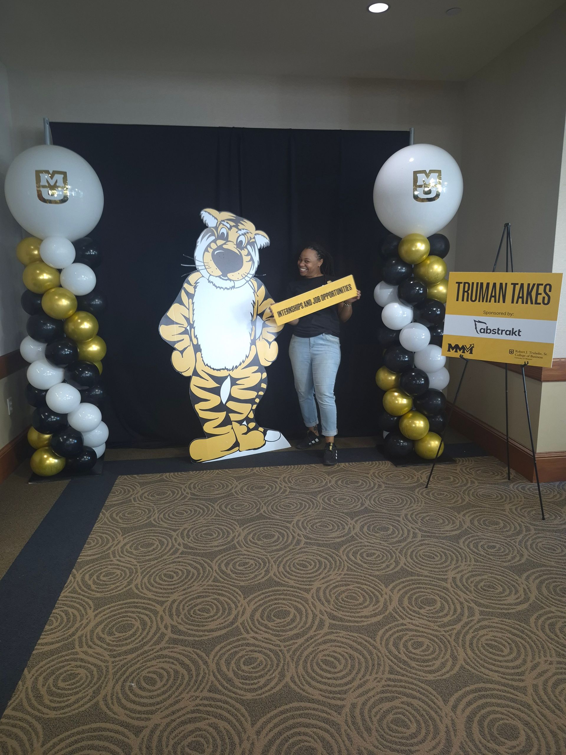 A person poses with a tiger cutout and balloons, in front of a backdrop, at a career fair.