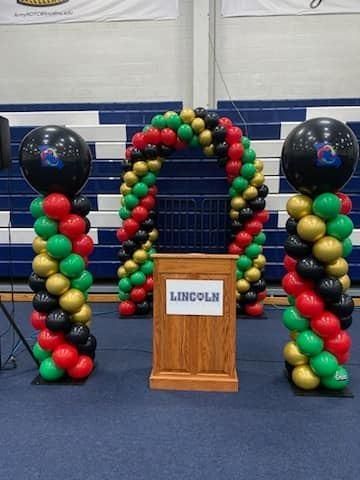 Balloon arch and columns with podium, in a gymnasium. Colors: black, red, green, gold.