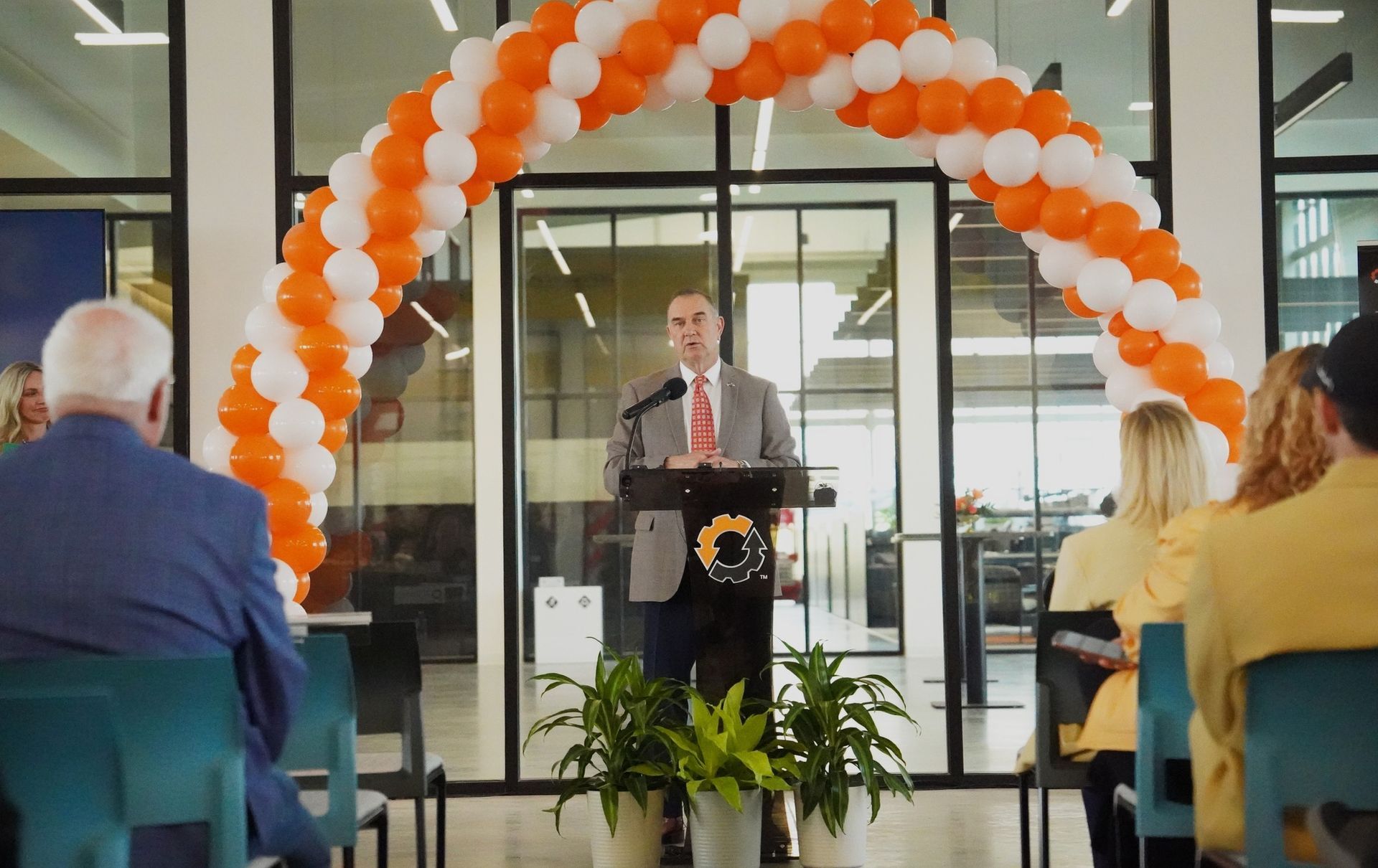 Man speaking at podium under orange and white balloon arch, at event.