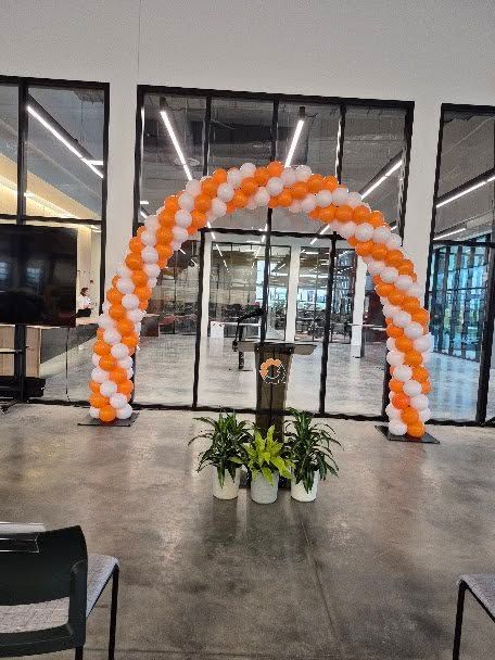 Orange and white balloon arch at an event entrance, with plants and podium in front of glass doors.