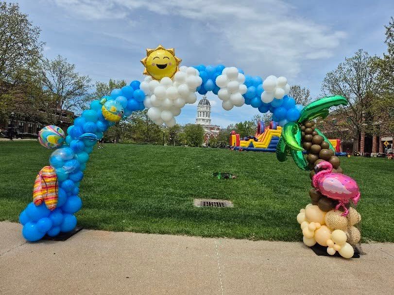Balloon arch with blue sky, sun, clouds, palm tree, and a flamingo. In a park setting.