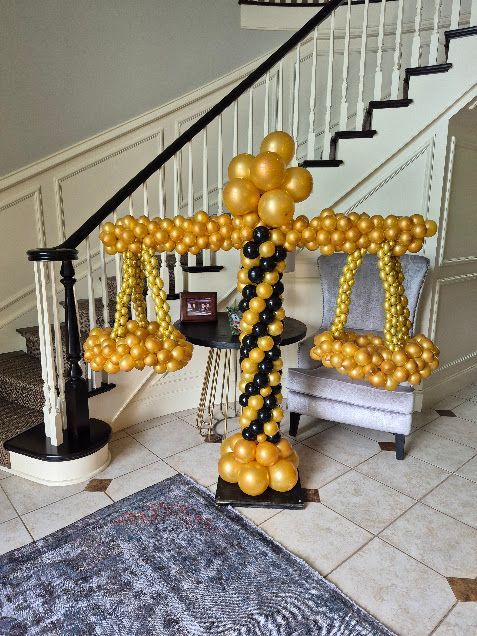 Gold and black balloon scales of justice sculpture, displayed indoors near a staircase and chair.