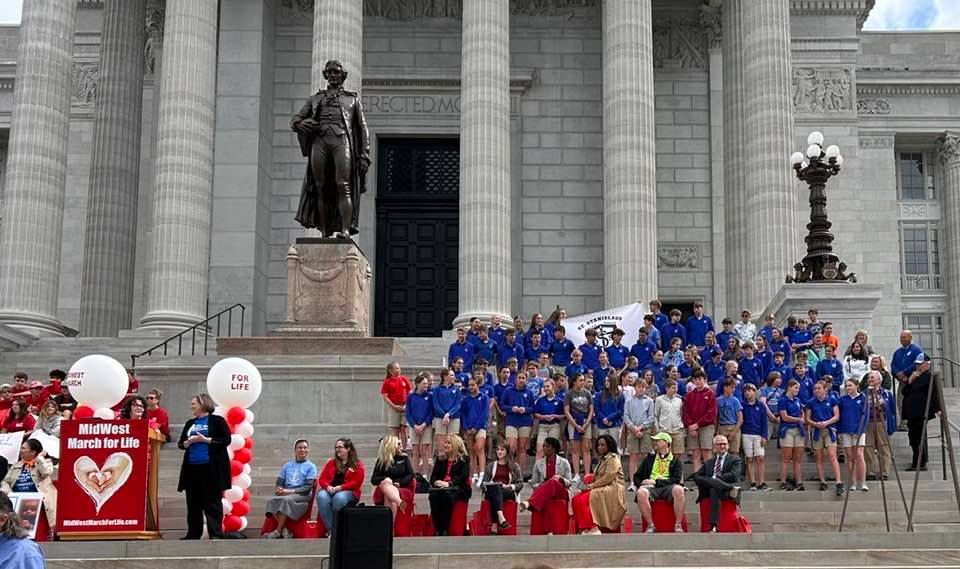 Group of people on steps of a building with a statue, holding signs. Red and blue clothing.