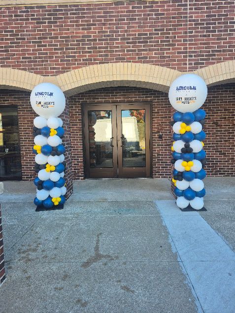 Two balloon columns flank a door. White, blue, and yellow balloons support a large white balloon with text.