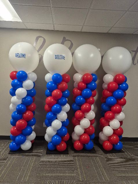 Four red, white, and blue balloon columns topped with white balloons, each with a logo, stand on black bases indoors.
