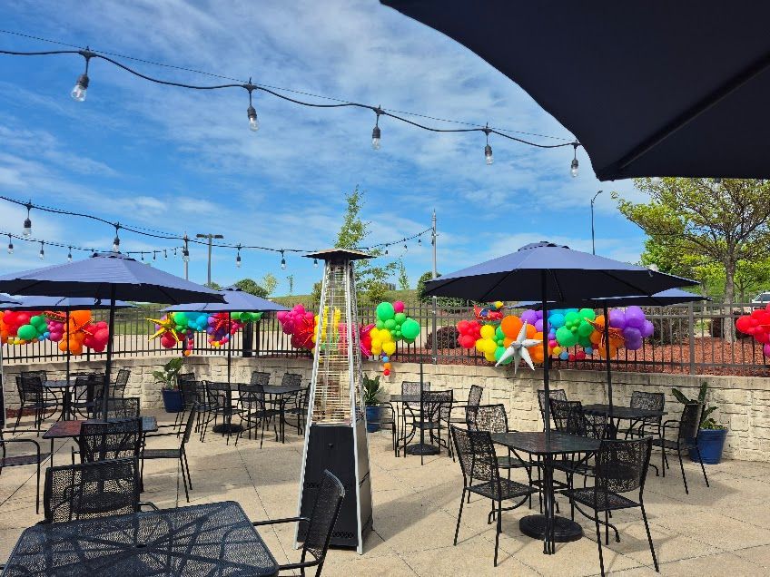 Outdoor restaurant patio with tables, umbrellas, string lights, and colorful balloons. Bright blue sky.
