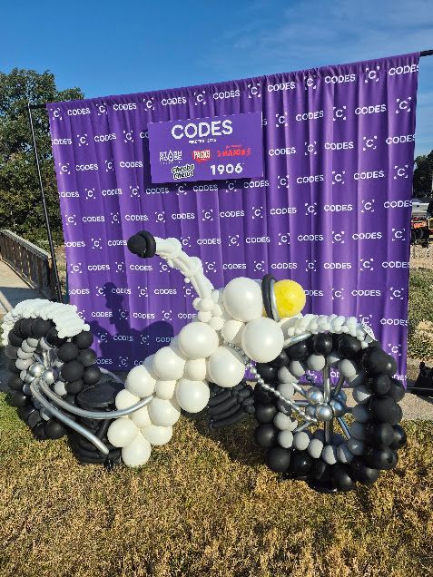 Balloon motorcycle sculpture in front of a purple banner with event branding.
