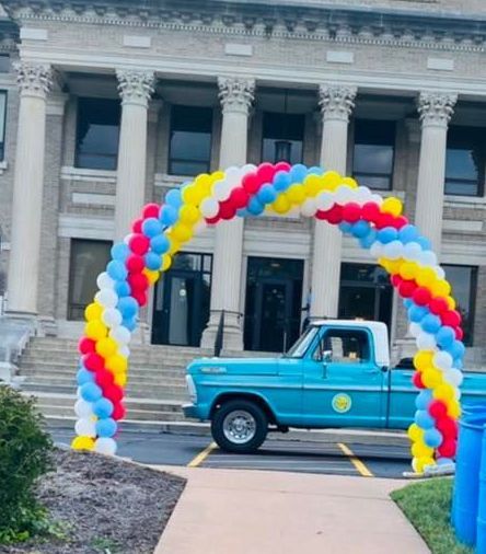 A blue truck is parked under a balloon arch in front of a building