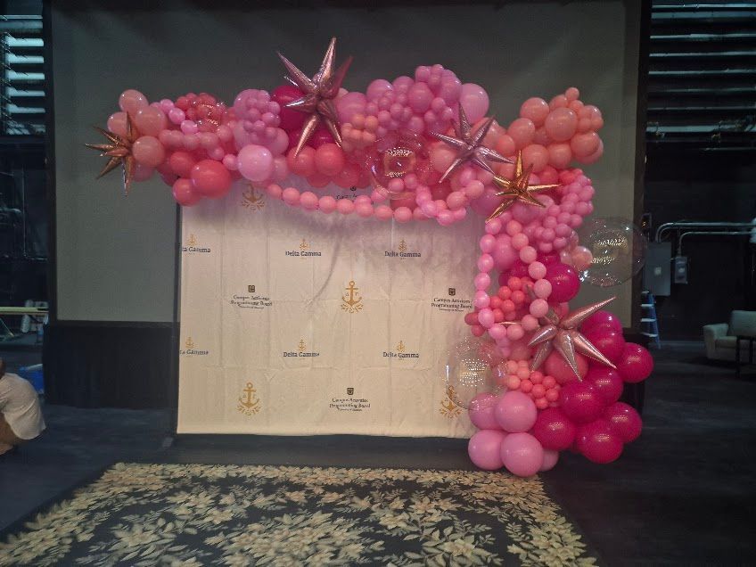 Pink and coral balloon arch with star accents, framing a white backdrop with text.
