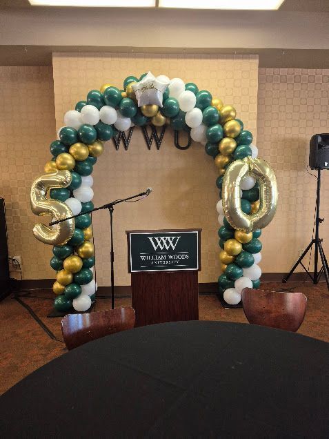 A decorated arch made of green, white, and gold balloons for a 50th celebration, with stage equipment.