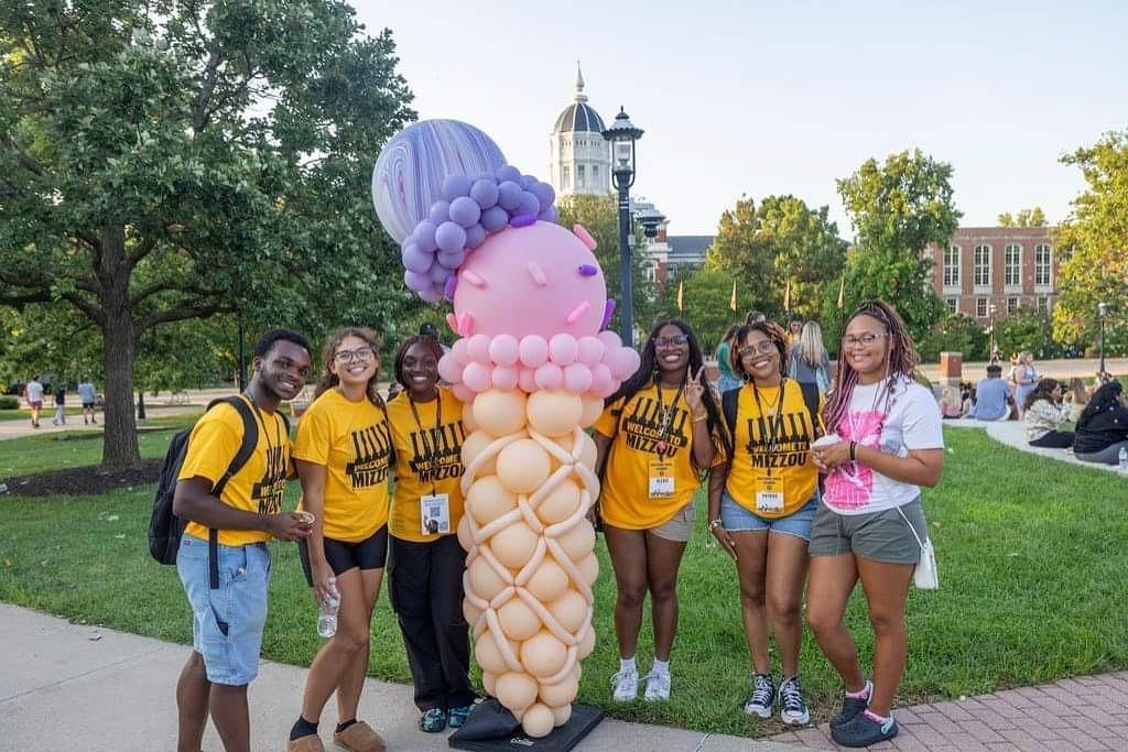 Group of people in yellow shirts posing with a large balloon ice cream cone on a campus green.