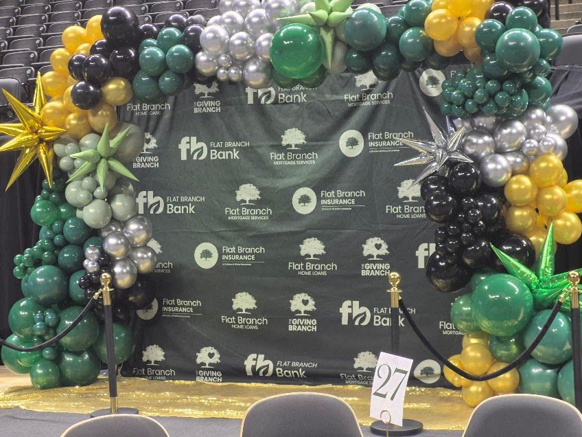 Green, black, and gold balloon arch over a backdrop with the FB bank logo at an event with tables.