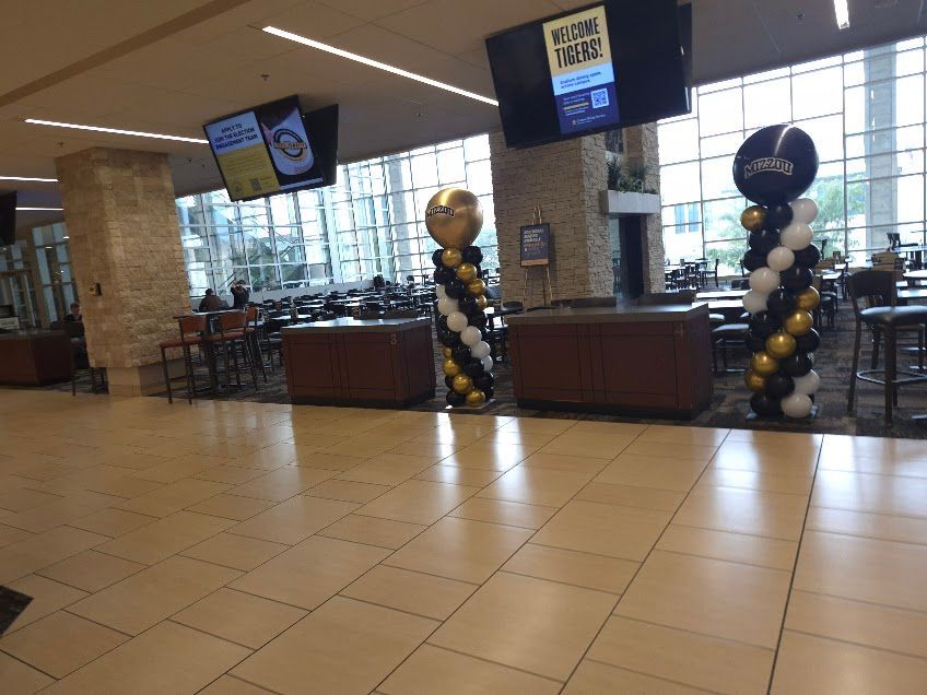 Interior view of a venue with balloon decorations. Tables, bar stools, TVs, and large windows are present.