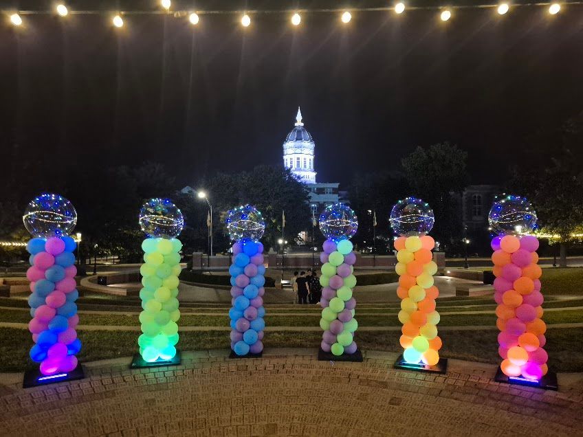 Six illuminated balloon pillars with disco balls, lit at night in front of a building with a dome.