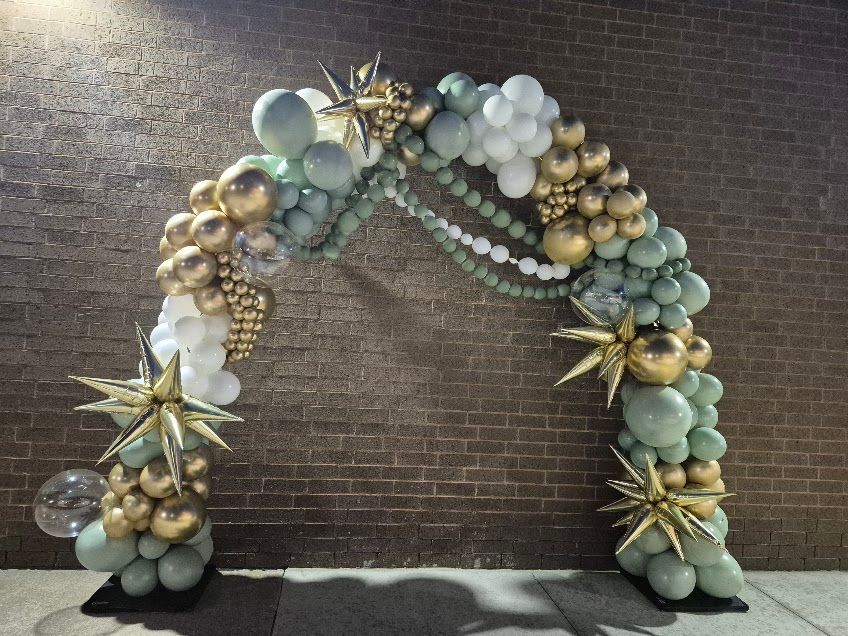 Balloon arch with gold, green, and white balloons, and star accents in front of a brick wall.
