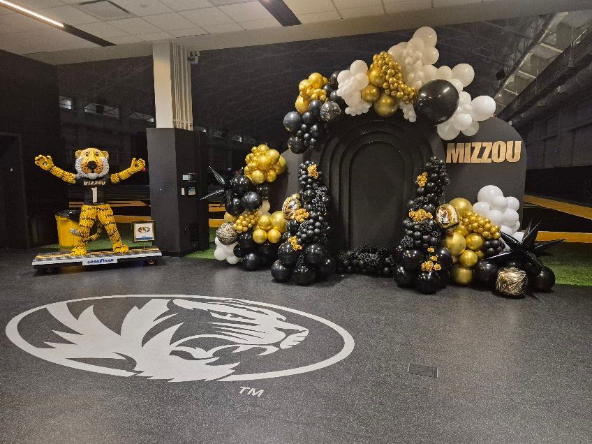 Mizzou-themed party: a tiger mascot, black and gold balloons, and a backdrop with “Mizzou” written on it.