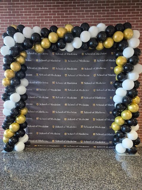 Black, gold, and white balloon archway with a backdrop, set against a brick wall.