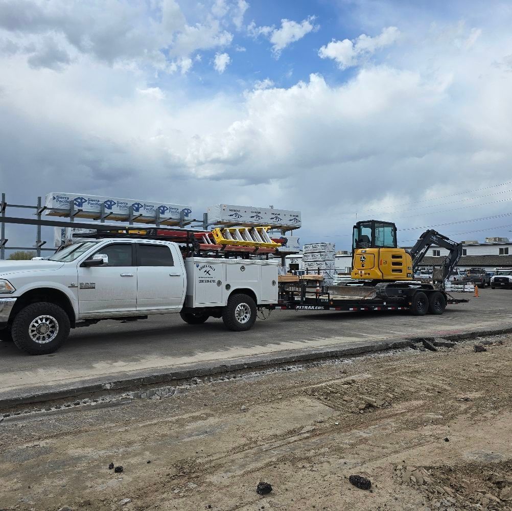 White truck towing a trailer with a yellow excavator on a construction site under a cloudy sky.