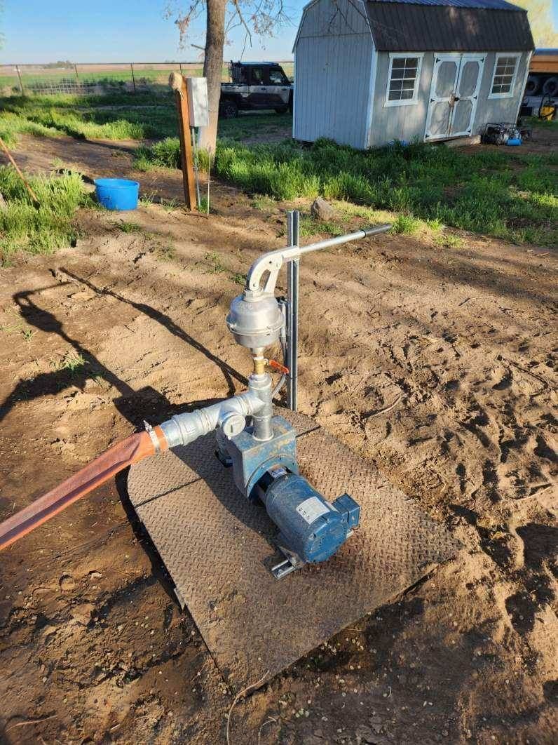 Well water pump system outdoors on a dirt surface near a shed and a blue barrel.