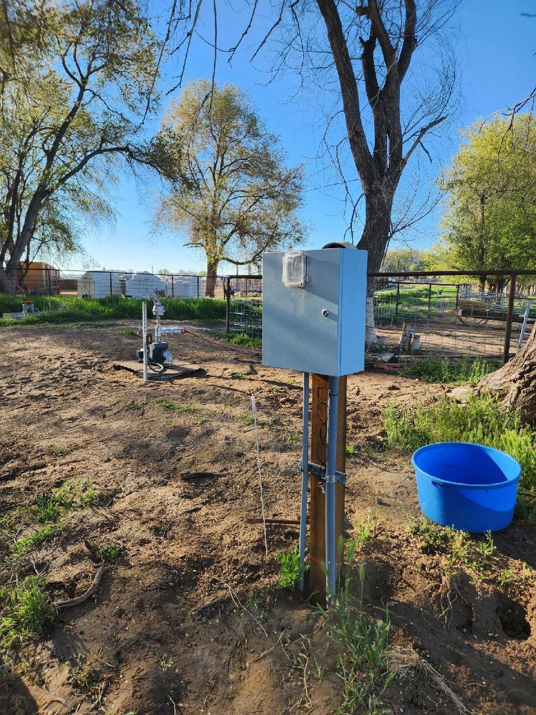 Outdoor electrical box on wooden post near a blue water trough, with trees and a clear sky in the background.