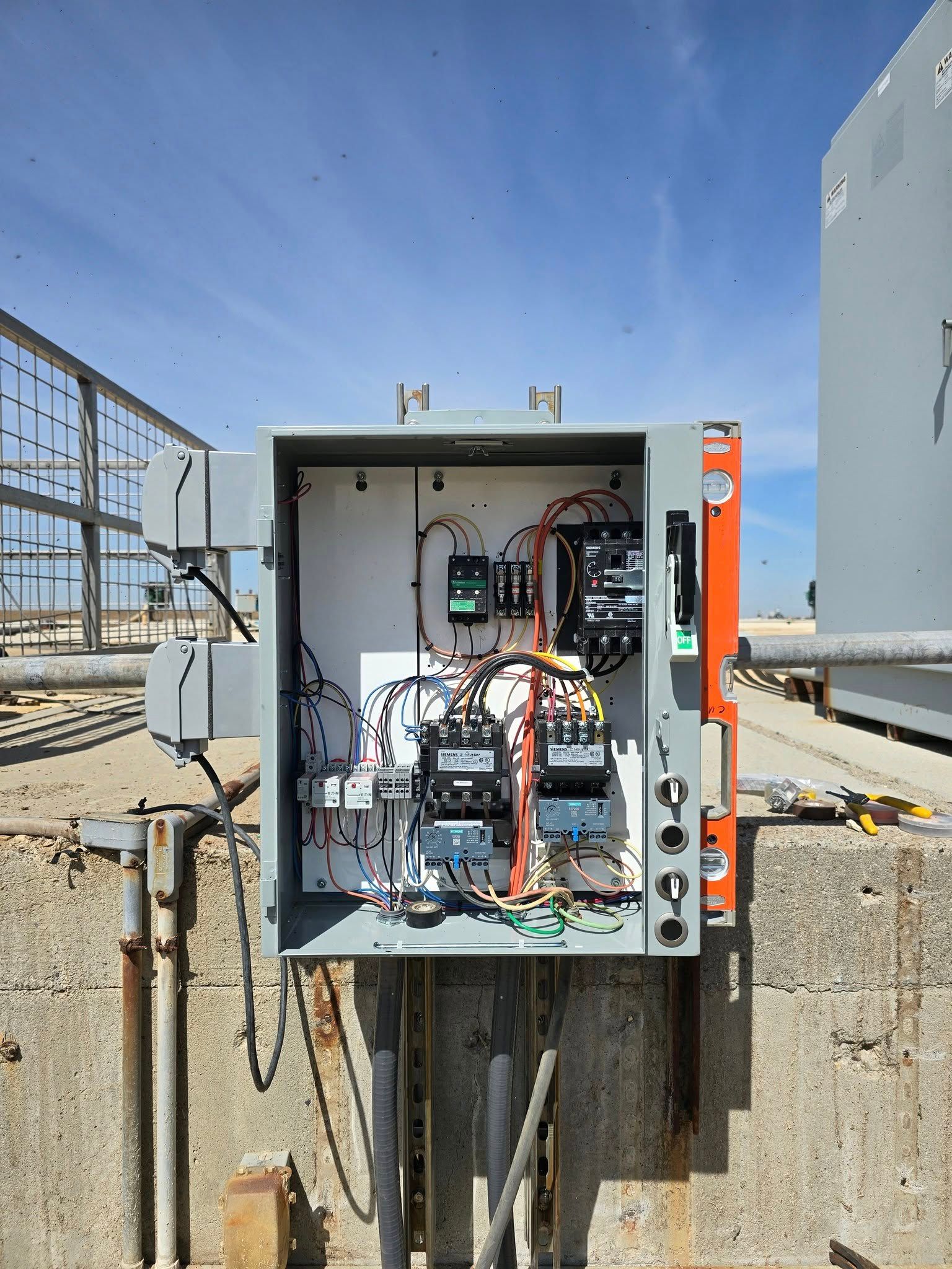 Electrical control box with open door, mounted outside. Wires, circuits, and components visible. Gray box on concrete.