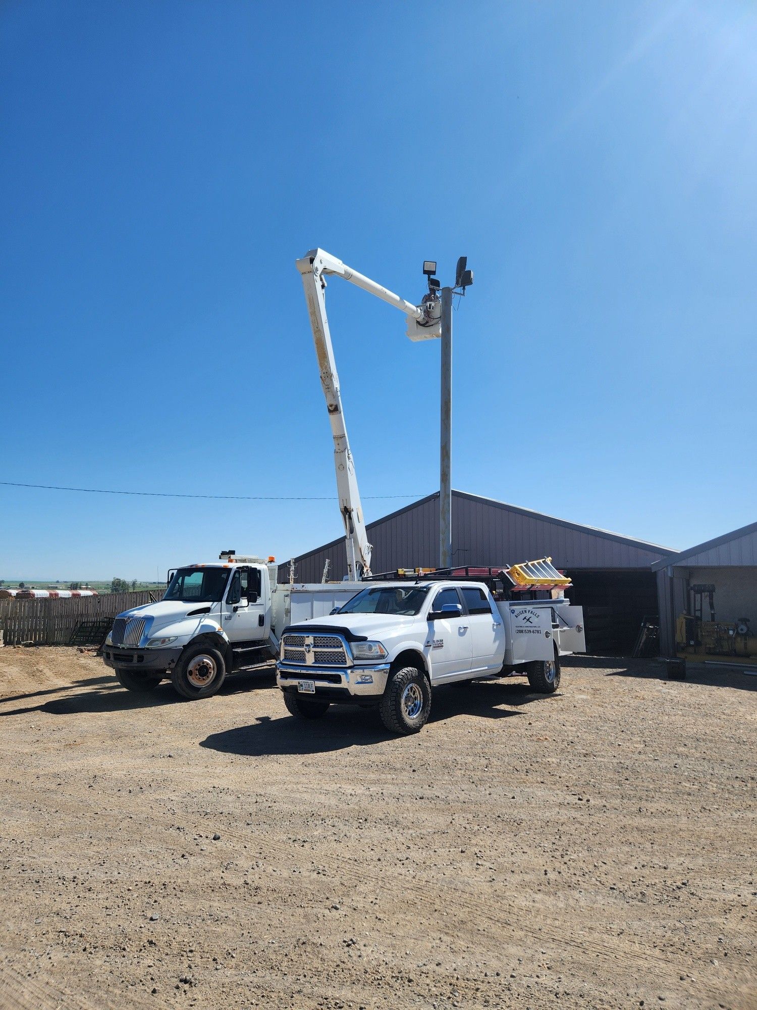 Two utility trucks parked near a tall pole with a raised bucket. Bright blue sky.
