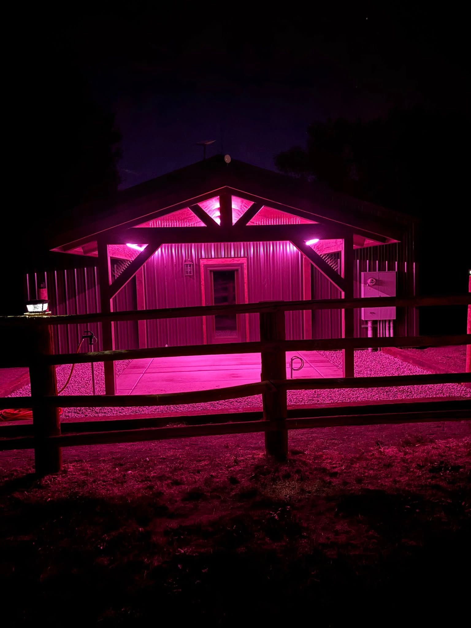 A wooden structure lit with bright pink lights at night. A wooden fence is in front of it.