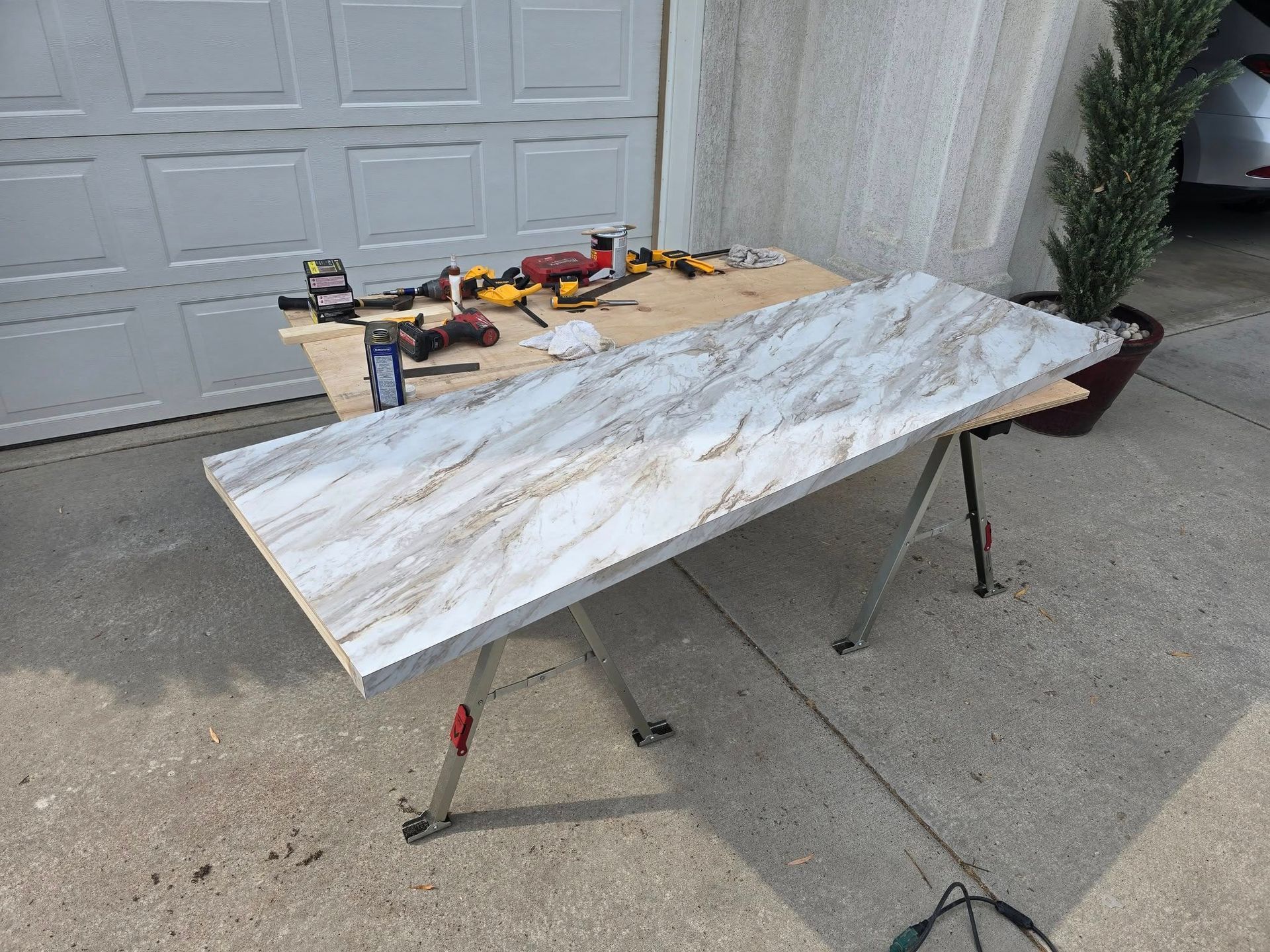 A large countertop sits on sawhorses outside a garage, with tools laid out on a wooden surface behind it.